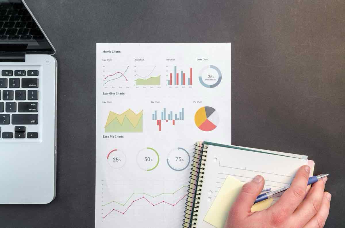 Hand with a pen reviewing printed charts next to a laptop and notebook